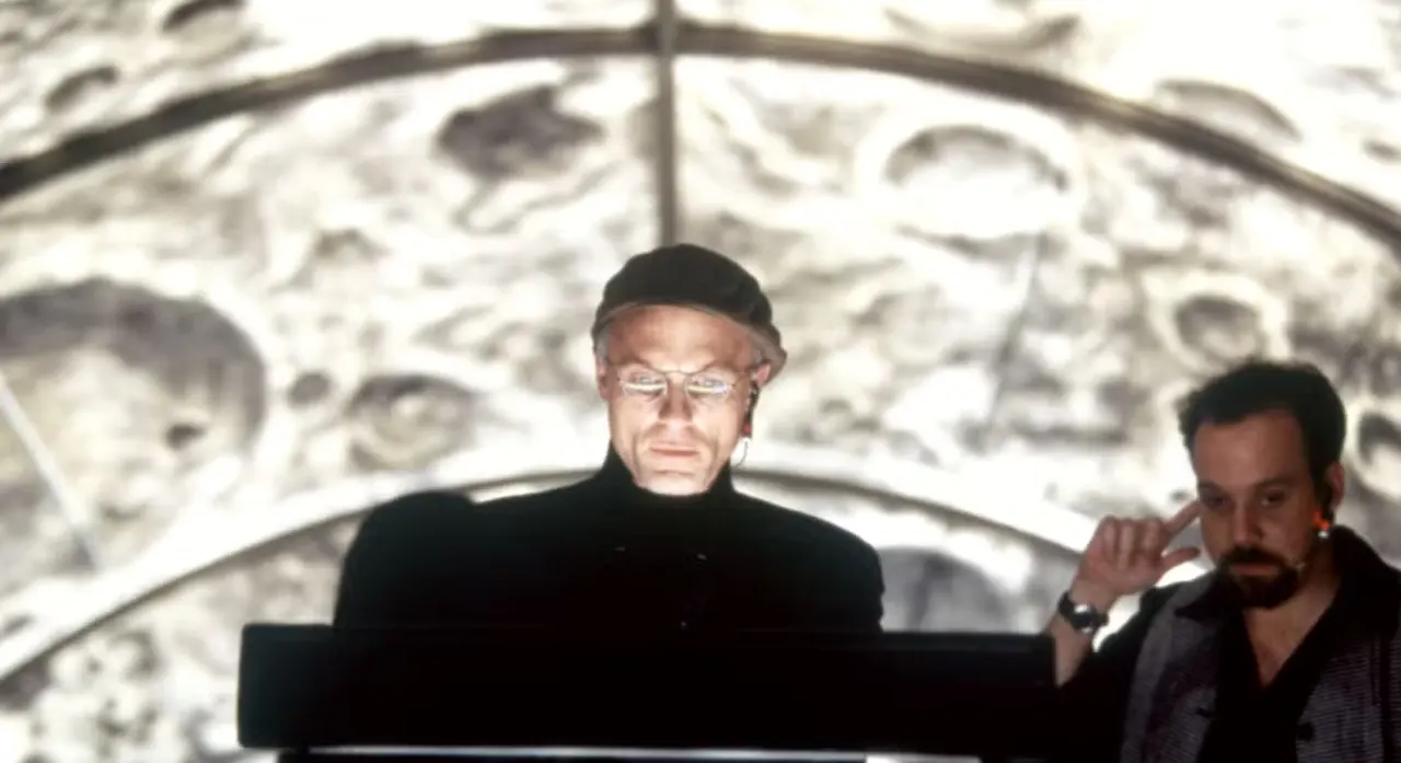 Ed Harris in a beret looks into a bright TV monitor; behind him are the craters of the moon