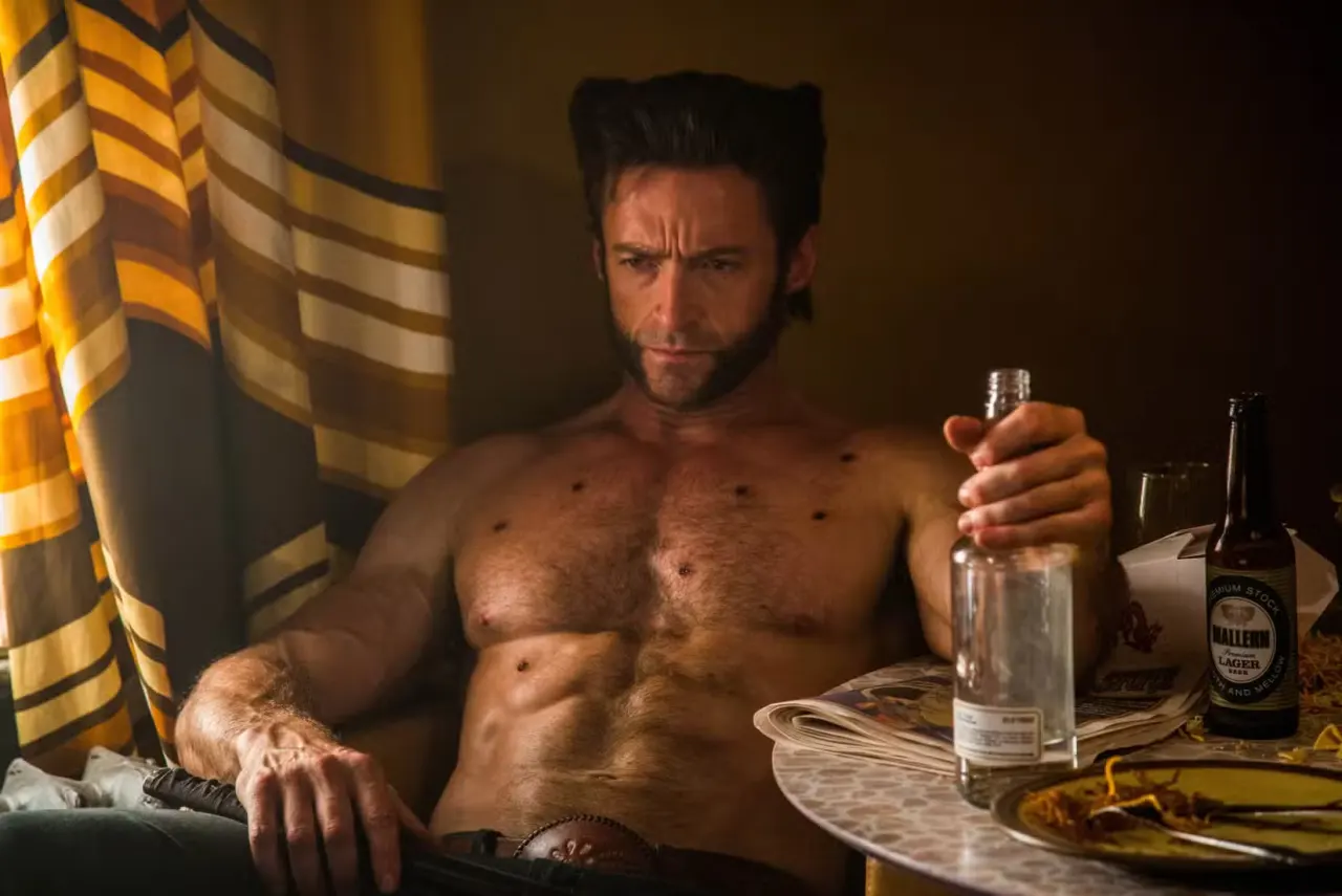 Wolverine, played by Hugh Jackman, sits in a chair. His expression is clearly irritated as he sits shirtless, one hand gripping a bottle of what’s probably alcohol.