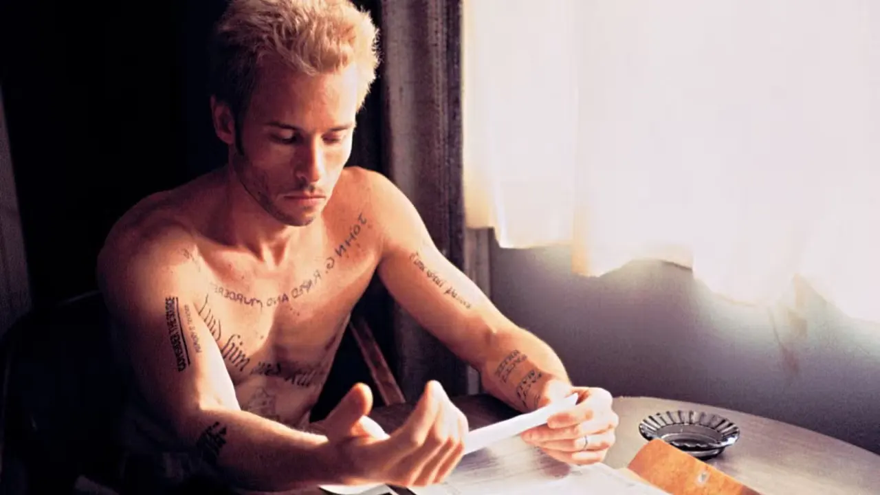 Leonard Shelby (Guy Peace) a shirtless young man with blonde hair covered in text tattoos, sits at a table in a hotel room and reads a note
